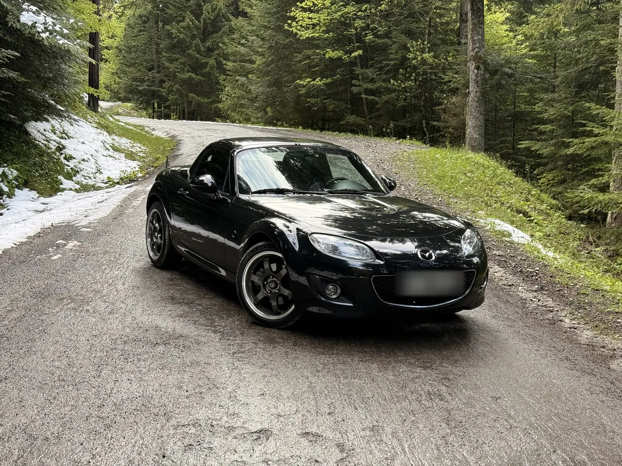 Mazda MX-5 NC noire sur une route forestière enneigée, toit fermé.